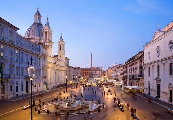Una piazza storica con una fontana al centro, circondata da architettura classica e un obelisco imponente.