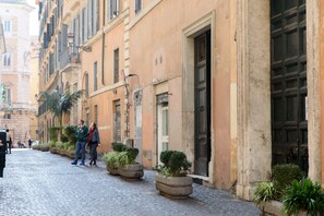 Una strada acciottolata fiancheggiata da edifici storici, con fioriere in pietra e piante verdi e un'ampia porta nera.