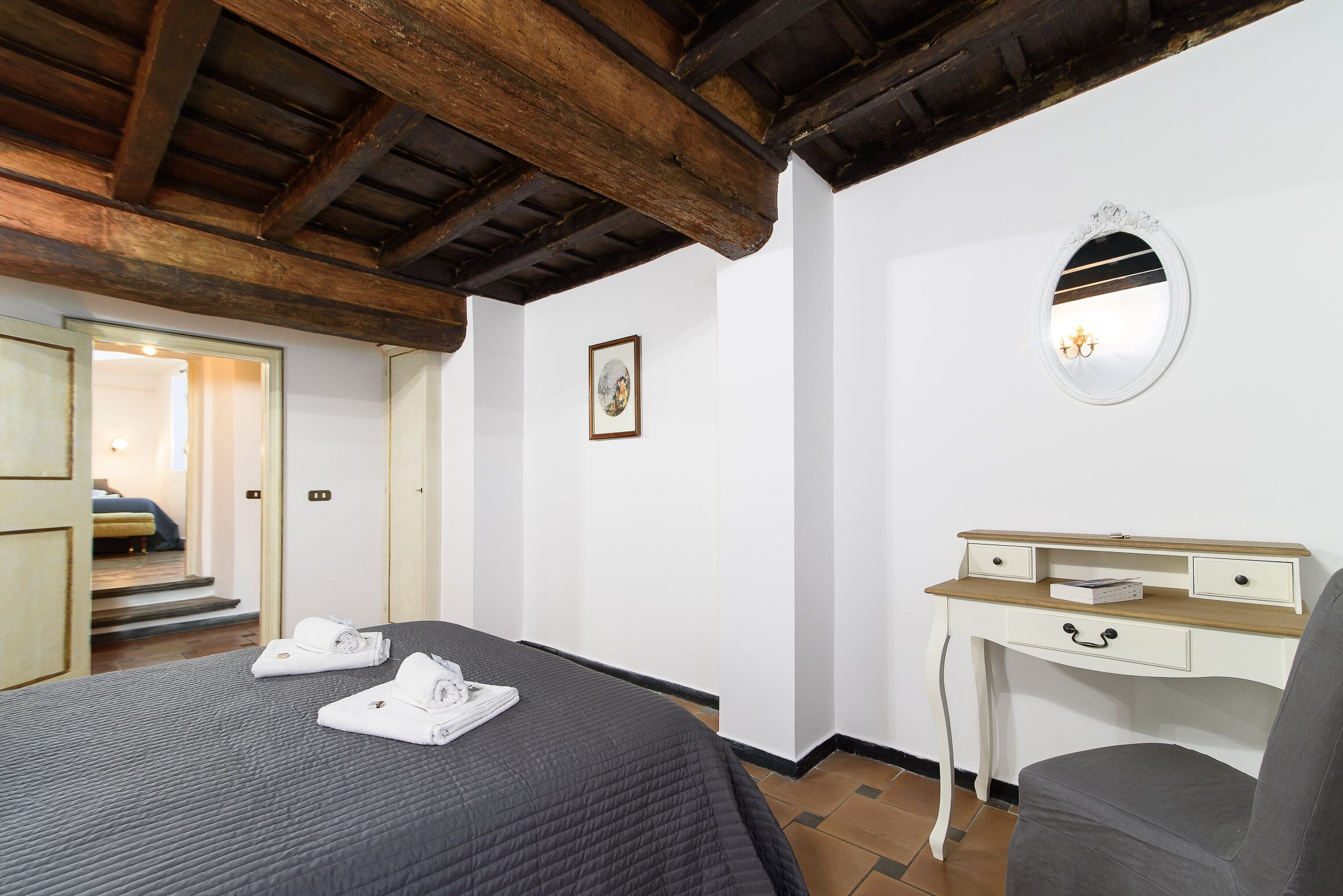 A bedroom with a bed, a desk, a chair, a mirror, and a door leading to another room.