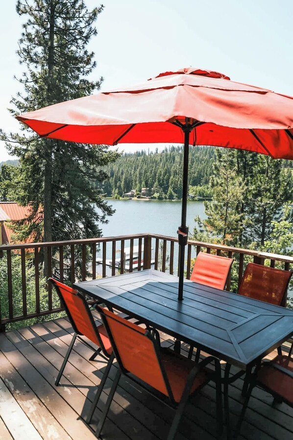 A wooden deck with a table and chairs, an umbrella, and a view of a lake and forest. - Lakefront Retreat - Private Dock & Beach! (Harrison)