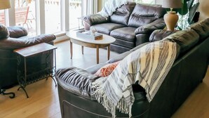 A spacious living room with a high wooden ceiling, large windows, a leather sofa, a coffee table, and a lamp.