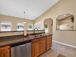 A modern kitchen with a dishwasher, wooden cabinets, a granite countertop, and a window with a view of greenery.
