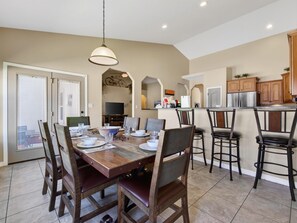 A dining area with a wooden table set for four, surrounded by chairs. A television is mounted on the wall, and a kitchen area is visible in the background.