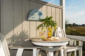 Una terraza de madera con una mesa redonda y dos sillas, una botella de aceite de oliva, una copa de vino y un bol de fruta.