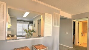 A hallway with a kitchen area, featuring a counter with stools, a window with blinds, and a door leading to another room. - Large Deck with Awesome Ocean & Marsh View! Free WiFi! Free Hunting Island pass! (St. Helena Island)