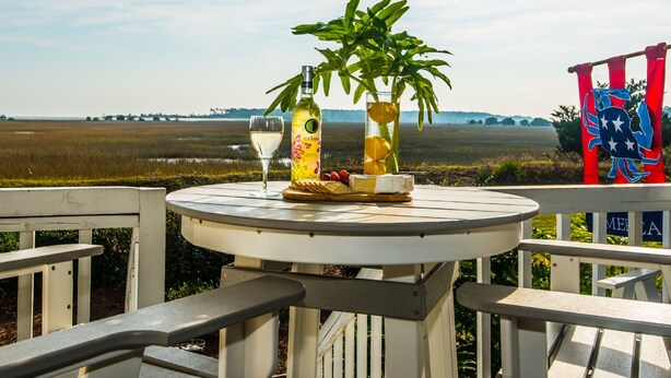 A table with a bottle of liquor, a glass, and a plant on a deck with a view of a field. - Large Deck with Awesome Ocean & Marsh View! Free WiFi! Free Hunting Island pass! (St. Helena Island)