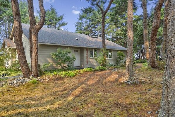 A single-story house with a sloped roof, surrounded by trees and a stone pathway leading to the front door.