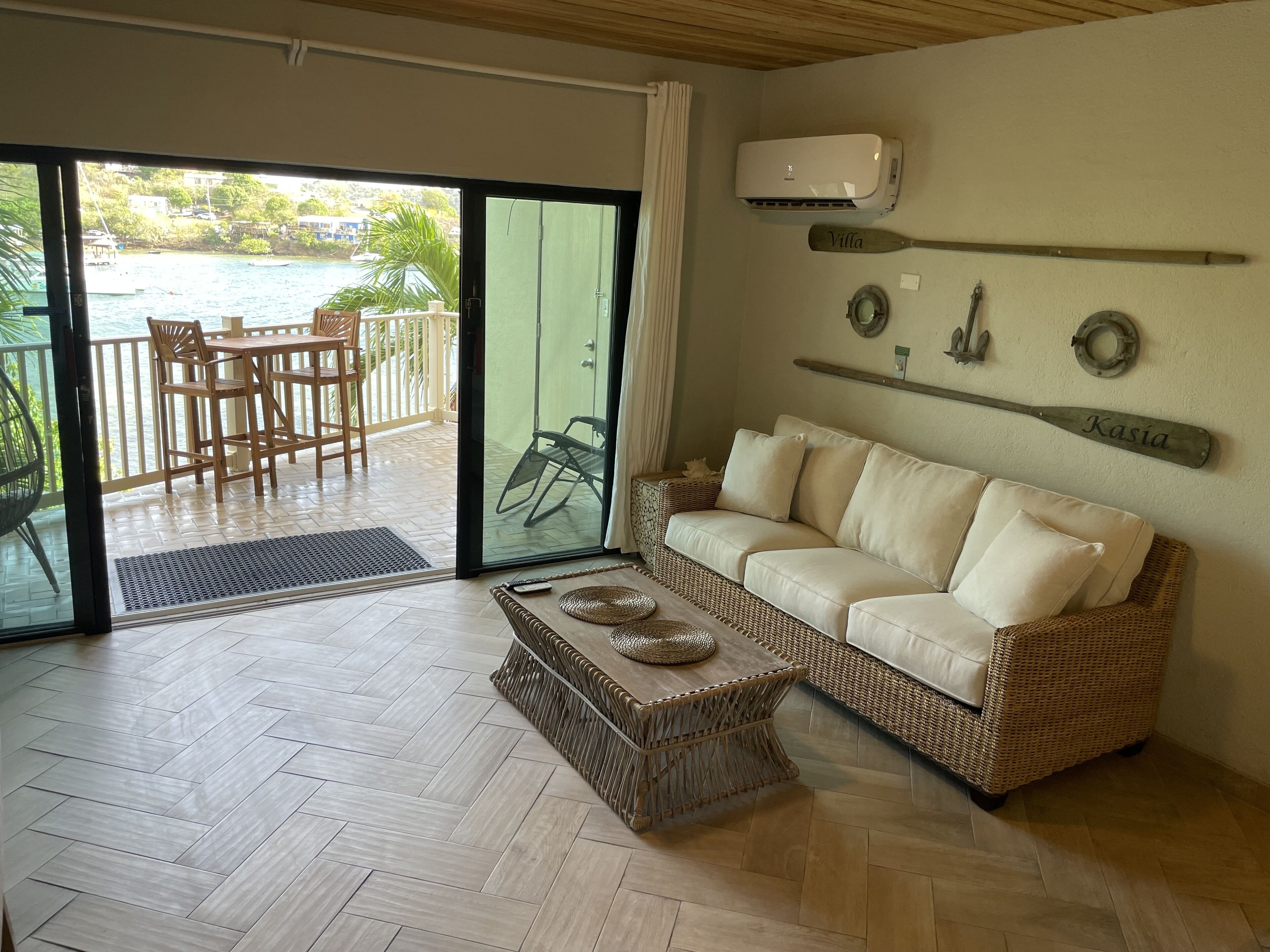 Una sala de estar con un sofá, una mesa de centro de mimbre, un ventanal de vidrio que da a un balcón con vista al mar y un aire acondicionado instalado.