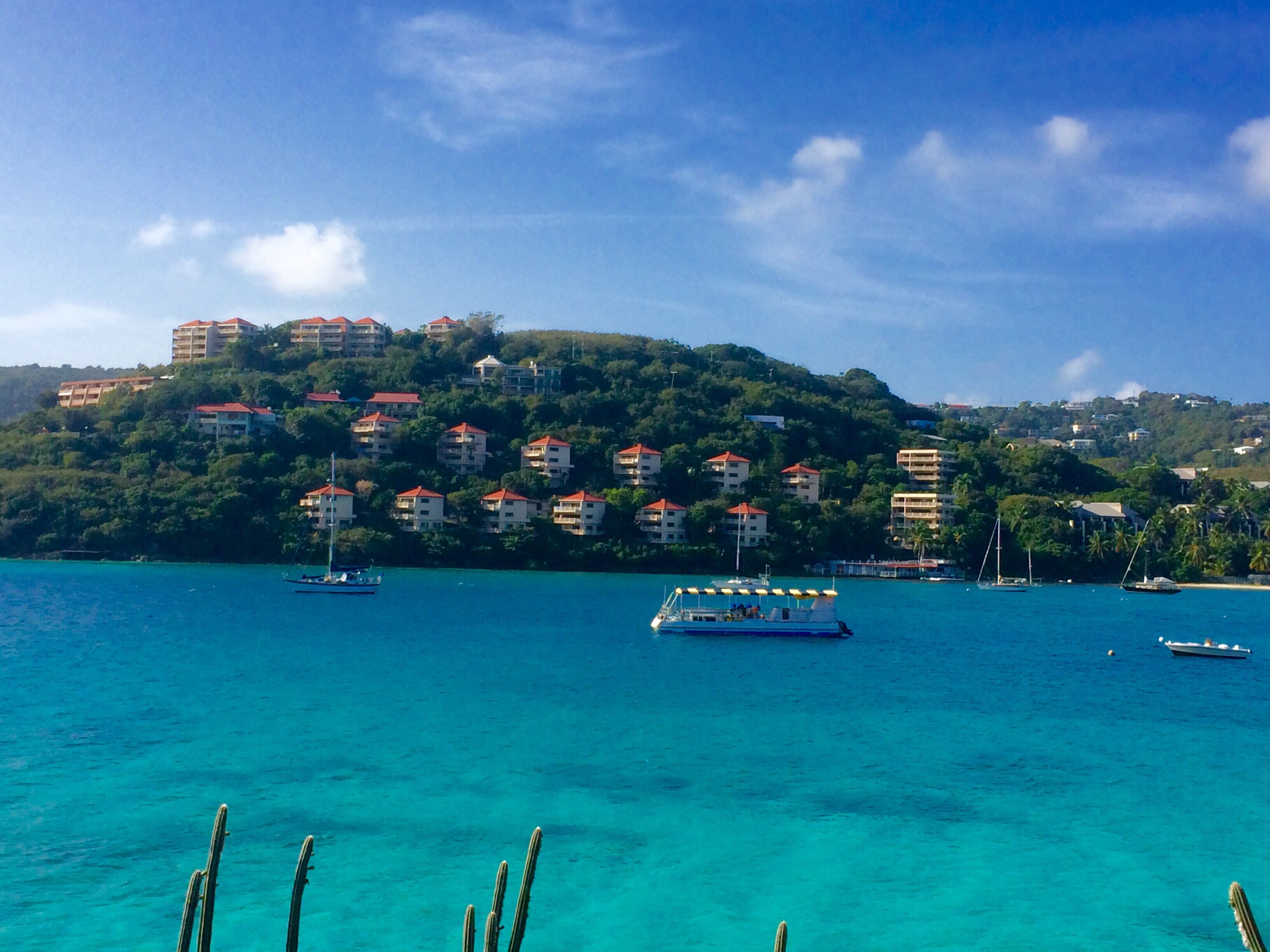 A coastal view with buildings on a hillside, clear turquoise water, and boats.