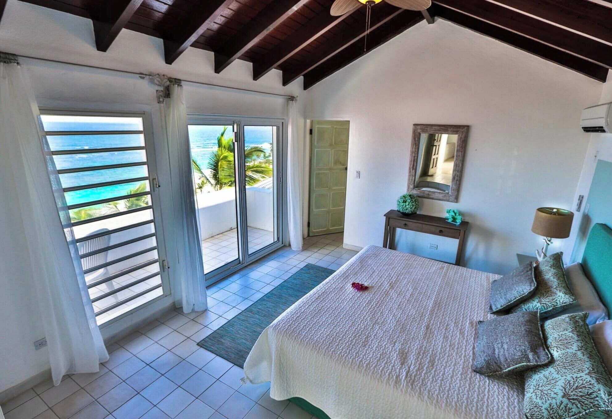 A bedroom with a bed, a nightstand, a mirror, and a view of the ocean through a sliding door.