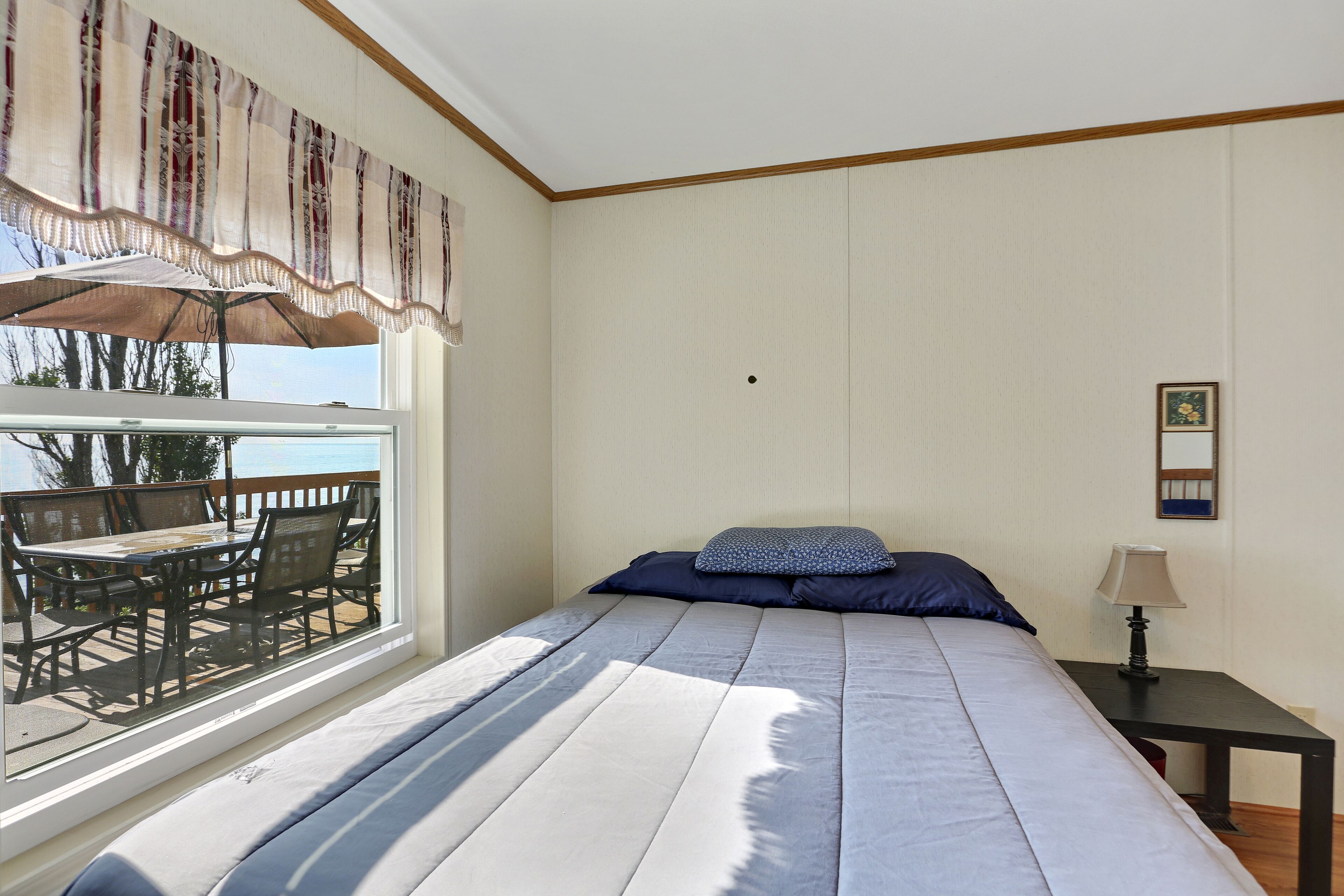 A bedroom with a large bed, a window with a view of the outdoors, a small table with a lamp, and a picture on the wall.