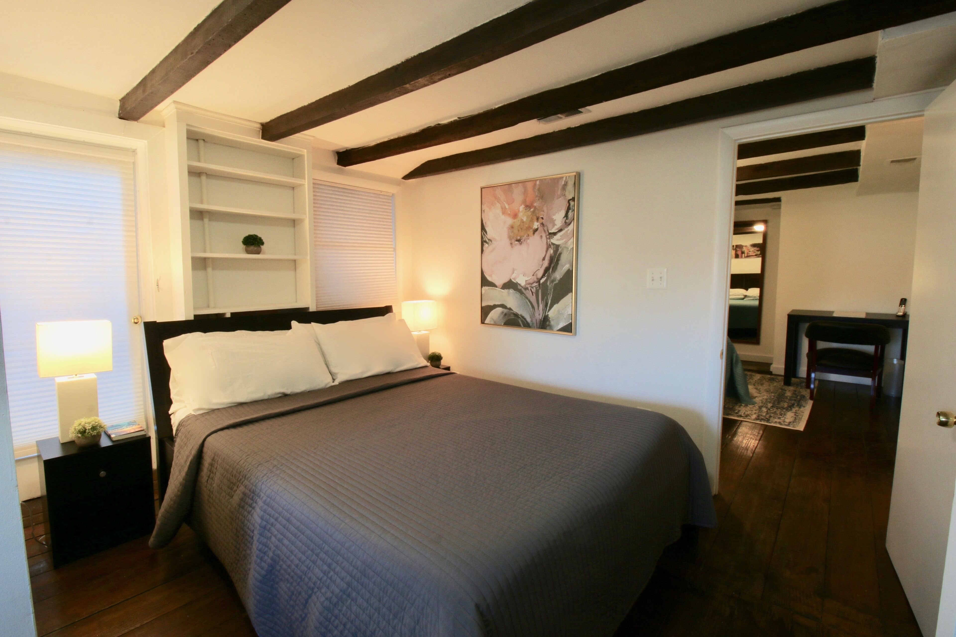 A bedroom with a bed, bedside tables, a lamp, a painting, and a wooden floor.
