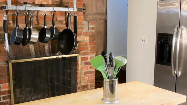 A kitchen with a brick wall, a wooden table, hanging pots, a glass vase with utensils, and a stainless steel refrigerator.