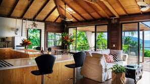 Una cocina moderna con techo de madera, zona de comedor con sofá blanco y sillas negras, y ventanas amplias que ofrecen vistas al exterior.