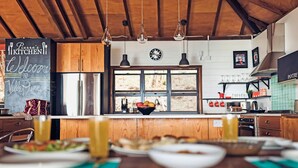 Une cuisine avec des meubles en bois, un réfrigérateur, un évier et une fenêtre donnant sur des arbres. On y trouve également des lampes suspendues et un panneau noir.