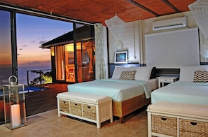 A bedroom with two beds, a nightstand, a bedside table, a lamp, and a view of the ocean.