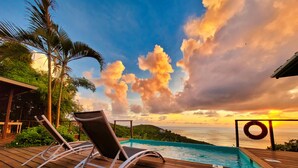 Une terrasse en bois avec des chaises longues et une piscine, offrant une vue panoramique sur l’océan et un magnifique coucher de soleil.