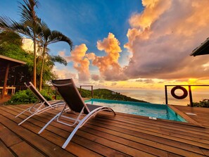 Eine Holzterrasse mit Liegestühlen und einem Pool mit Blick auf die Küste und einen Sonnenuntergang.