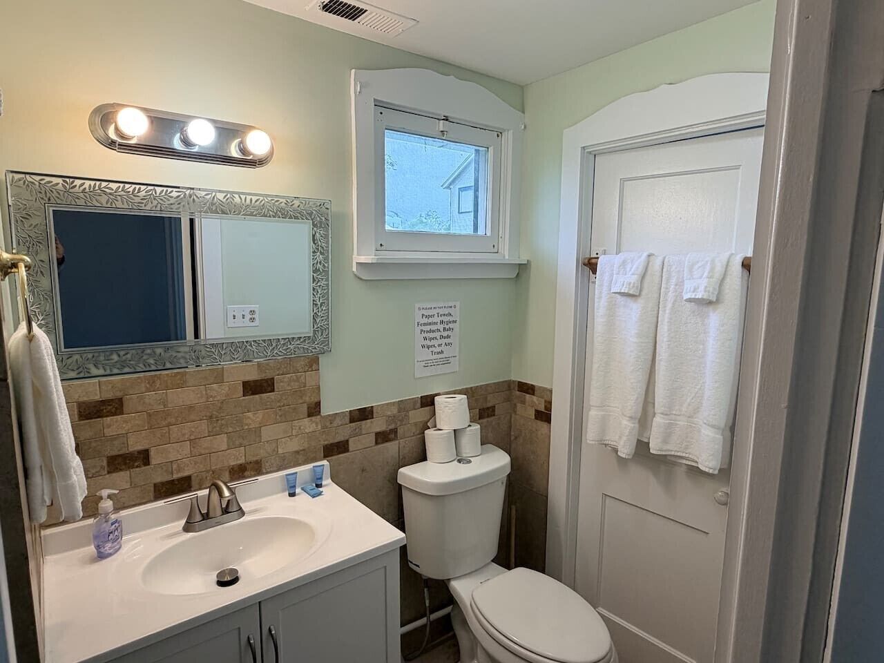 A bathroom with a sink, mirror, toilet, and a window.