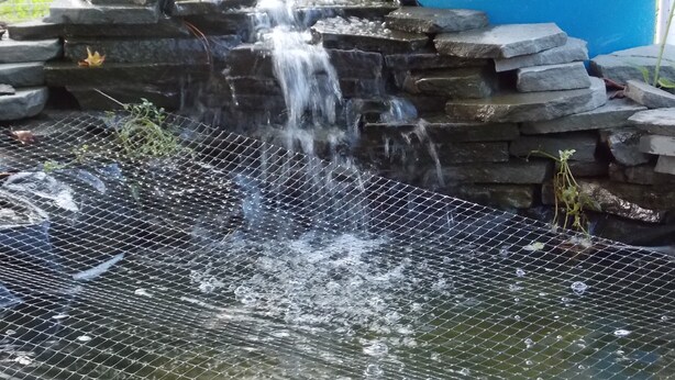 Ein kleiner Wasserfall mit einem Maschendrahtzaun davor, umgeben von Steinpodesten und einem blauen Fass.