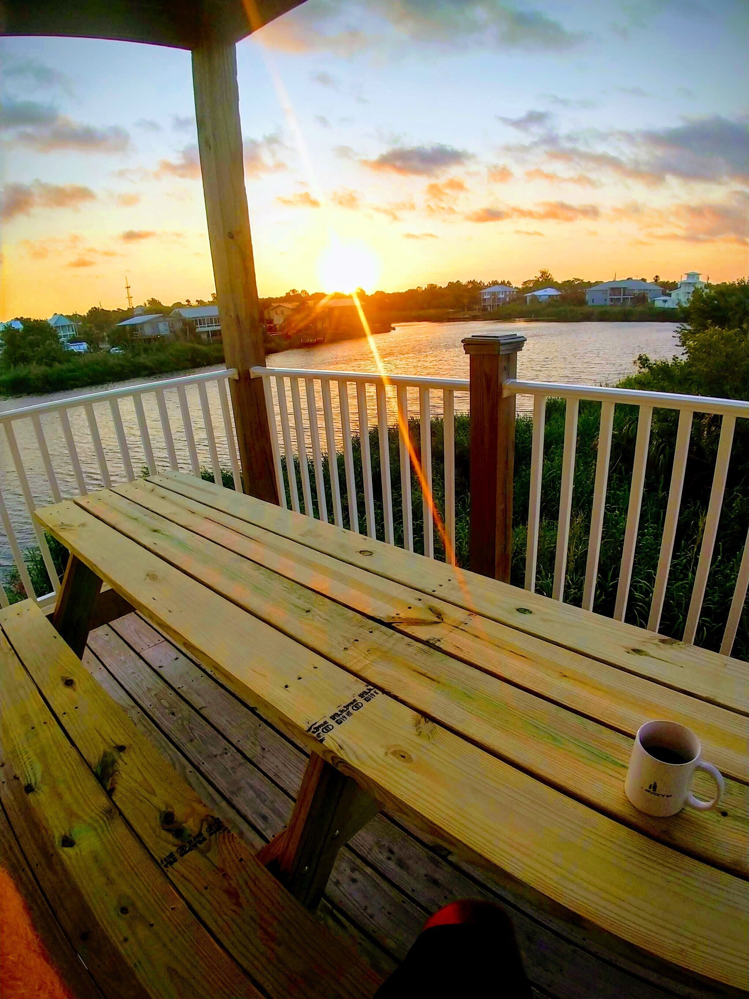 A wooden deck with a bench and a mug, overlooking a river with houses in the background during sunset.