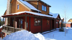 A two-story log cabin with a stone chimney, a covered porch, and a snow-covered roof.