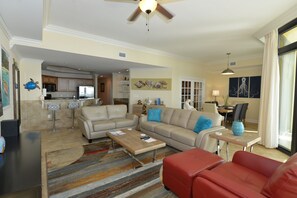 A spacious living room with a beige sofa, a red sofa, and a coffee table. There is a kitchen area in the background with a refrigerator, microwave, and cabinets.