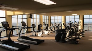 Ein Fitnessraum mit mehreren Laufbändern und stehenden Fahrrädern, die in Reihen angeordnet sind, mit großen Fenstern, die natürliches Licht und Blick auf die Stadt bieten.