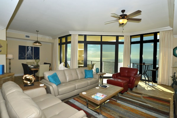 A spacious living room with a large window, a ceiling fan, and a dining area with a view of the ocean.