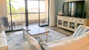 A spacious living room with a large sliding glass door leading to a balcony, a flat-screen TV, a white sofa, and a wooden coffee table. - Tradewind Mexico Beach 2/2 with balcony beach view (Mexico Beach)