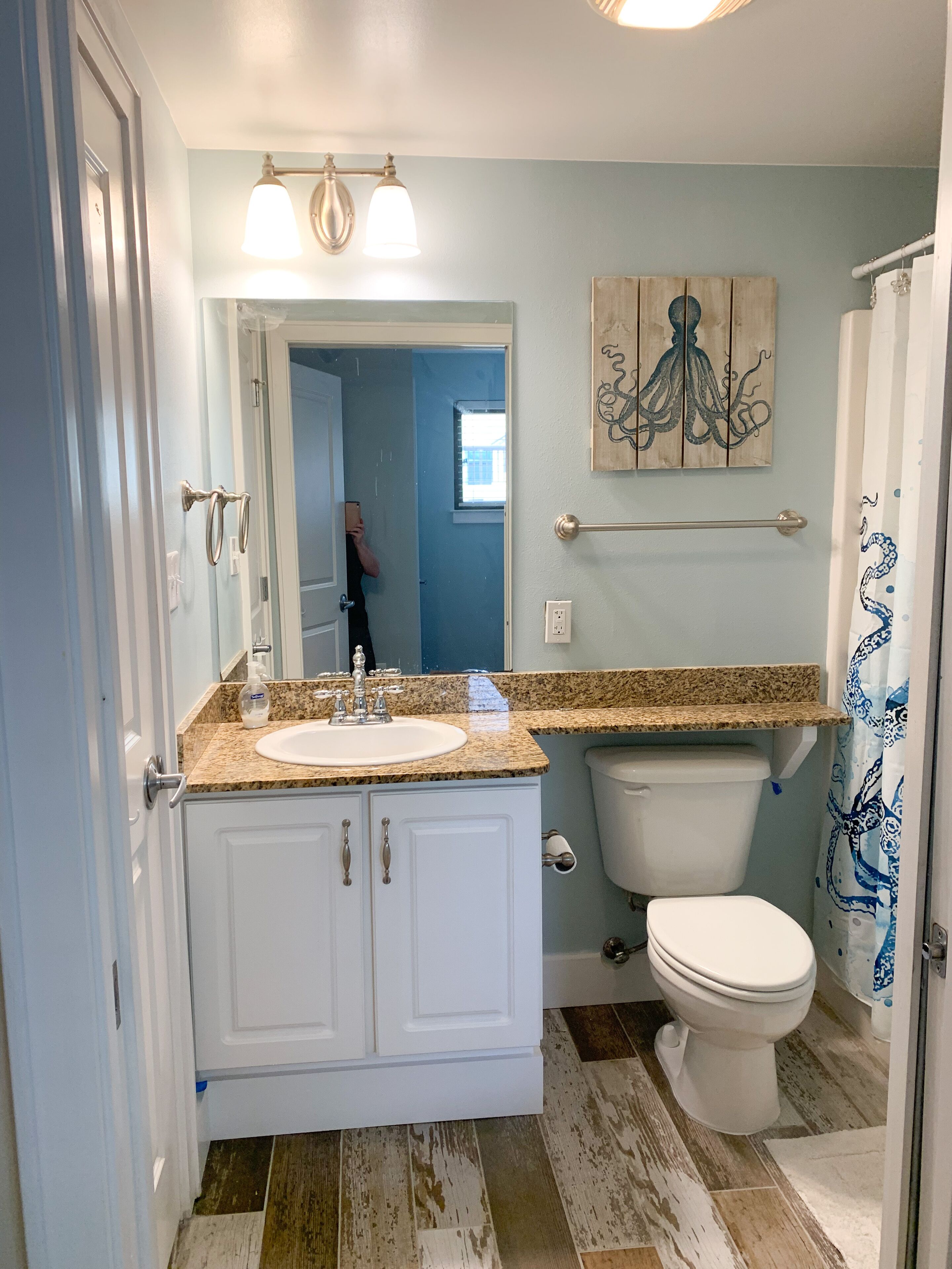 A bathroom with a white sink, vanity, and toilet. There is a wooden artwork above the toilet and a towel rack.