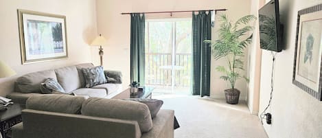 Een woonkamer met een bank, een lamp, een televisie, een plant en een raam met gordijnen.
