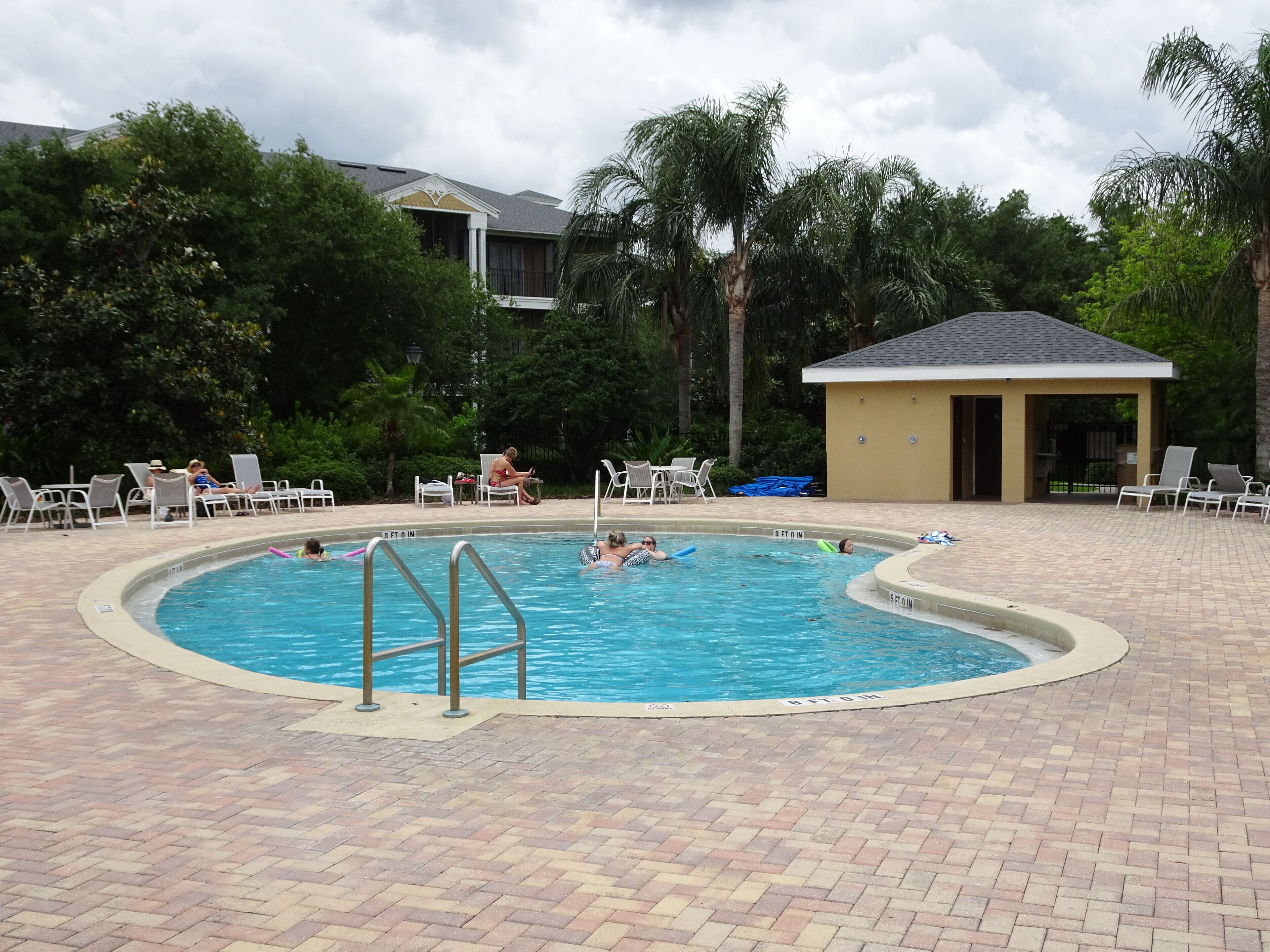 A swimming pool with a brick deck, surrounded by lounge chairs and palm trees.