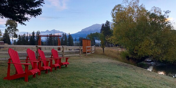 A grassy area with red Adirondack chairs, a wooden structure, and a view of mountains and a lake. - 90+ Acre Ranch Directly On The Elk River In The Heart of the Canadian Rockies! (Sparwood)