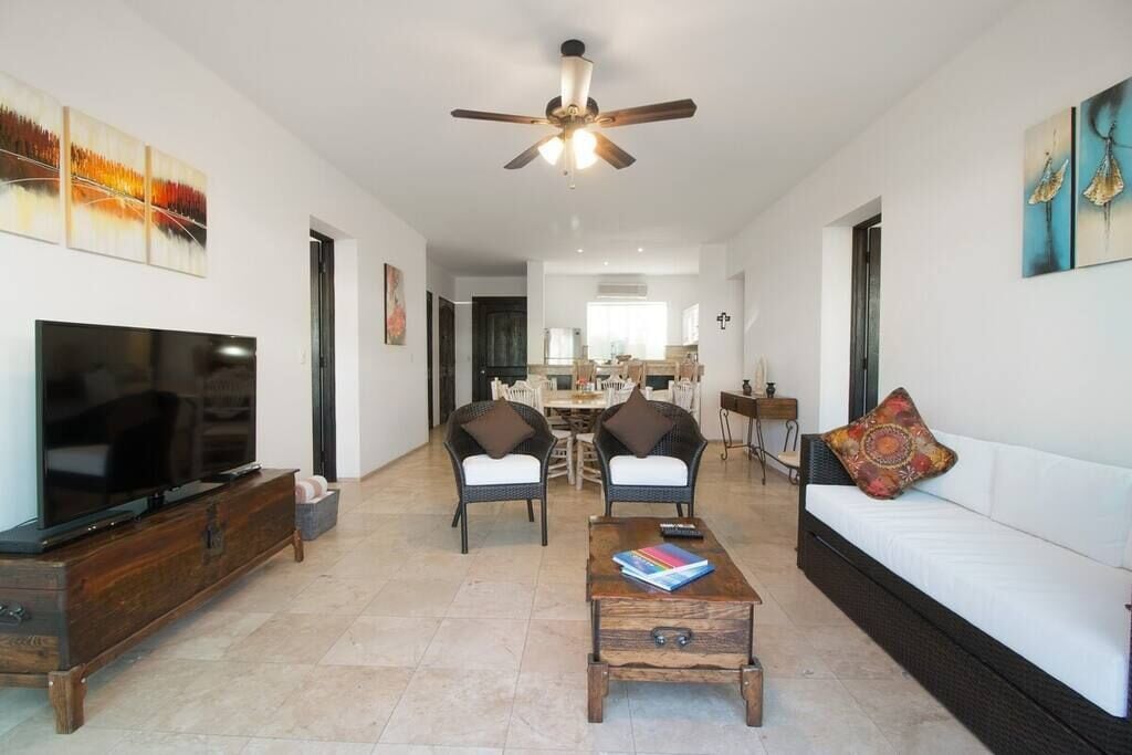 A spacious living room with a flat-screen TV, a ceiling fan, and a dining area in the background.