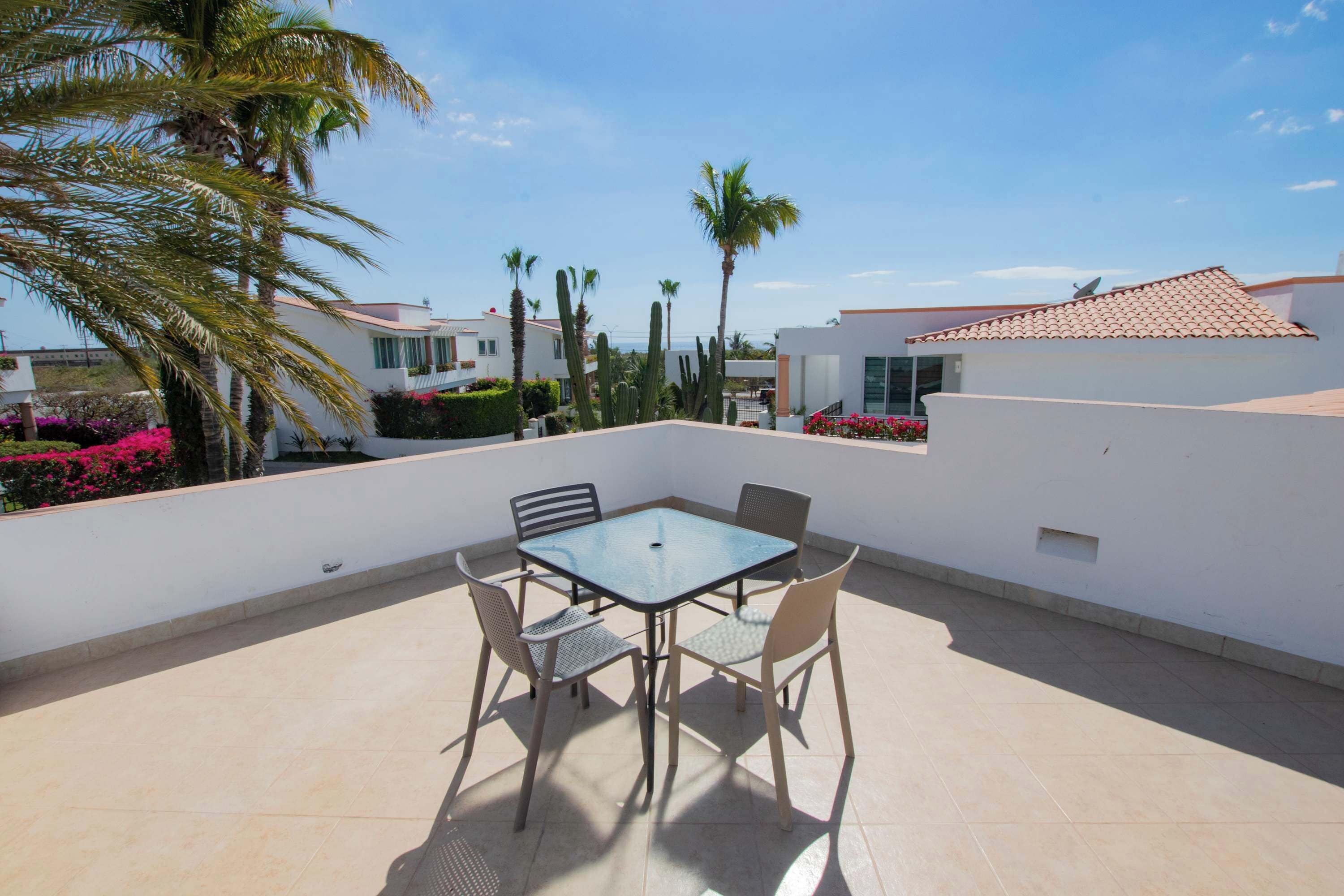 A patio with a glass table and four chairs, surrounded by palm trees and cacti, with a view of other buildings in the background.