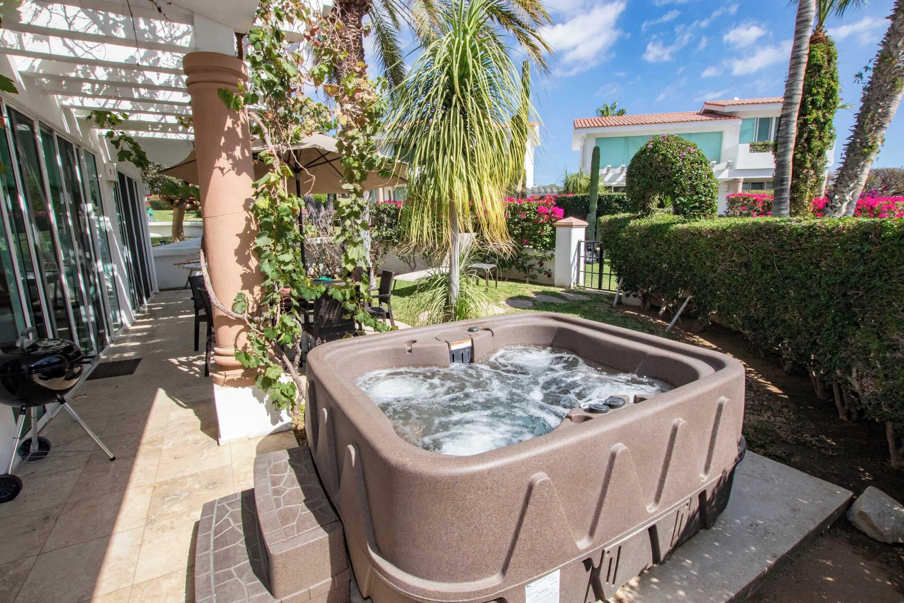 Un jacuzzi en un jardín con una pérgola, palmeras y un edificio al fondo.