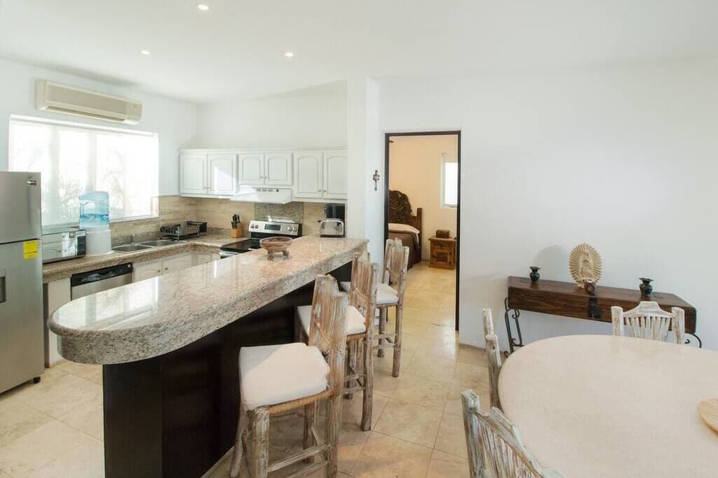 Una cocina con encimera de granito, armarios blancos, un comedor con mesa y sillas de madera, y vista a otra habitación.