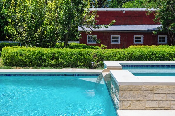 A swimming pool with a stone border, a red building with white trim in the background, and well-maintained greenery.