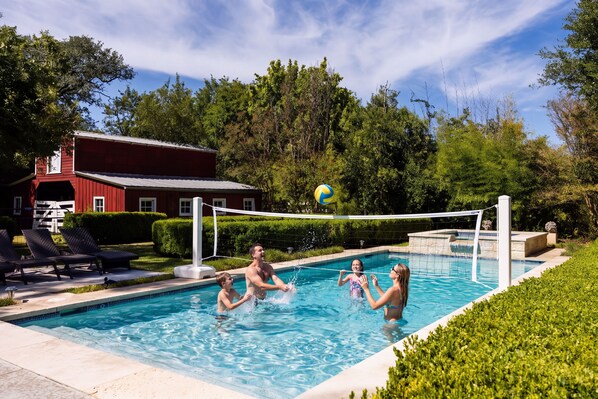 Una piscina con red de voleibol, sillas de descanso y un edificio rojo al fondo.