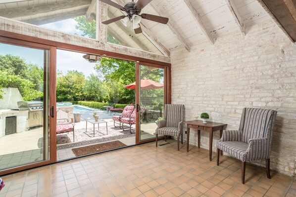 A spacious patio with a glass door leading to a pool area, a ceiling fan, and a brick wall.