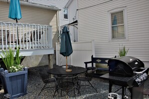 A patio with a round table, chairs, a grill, and a blue umbrella.