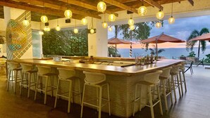 Eine Bar mit einer langen Theke, mehreren Sitzhocker, hängenden Lampen und Meerblick.