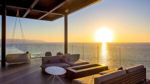 Ein moderner Außenbereich mit Sitzgelegenheiten und Blick auf den Sonnenuntergang über dem Meer, ausgestattet mit einer Hängematte, Sofas und einem kleinen Tisch.