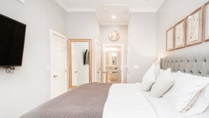 A bedroom with a large bed, a flat-screen TV mounted on the wall, and a hallway leading to a bathroom.