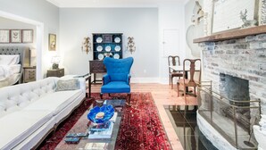 A living room with a white sofa, a blue armchair, a glass coffee table, a red patterned rug, a fireplace, and a wall-mounted shelf with decorative plates.