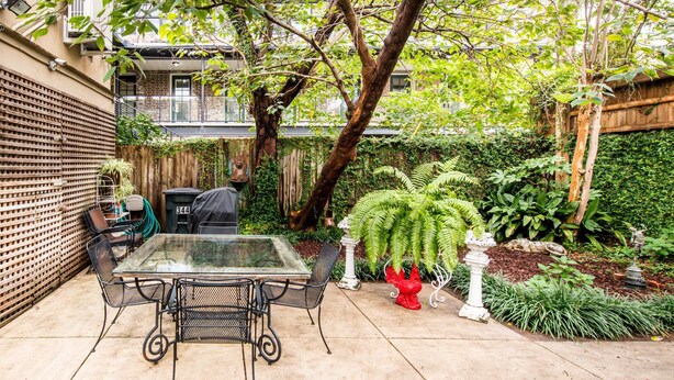 A glass-top table with four chairs in a garden area with a wooden fence, potted plants, and a small statue.