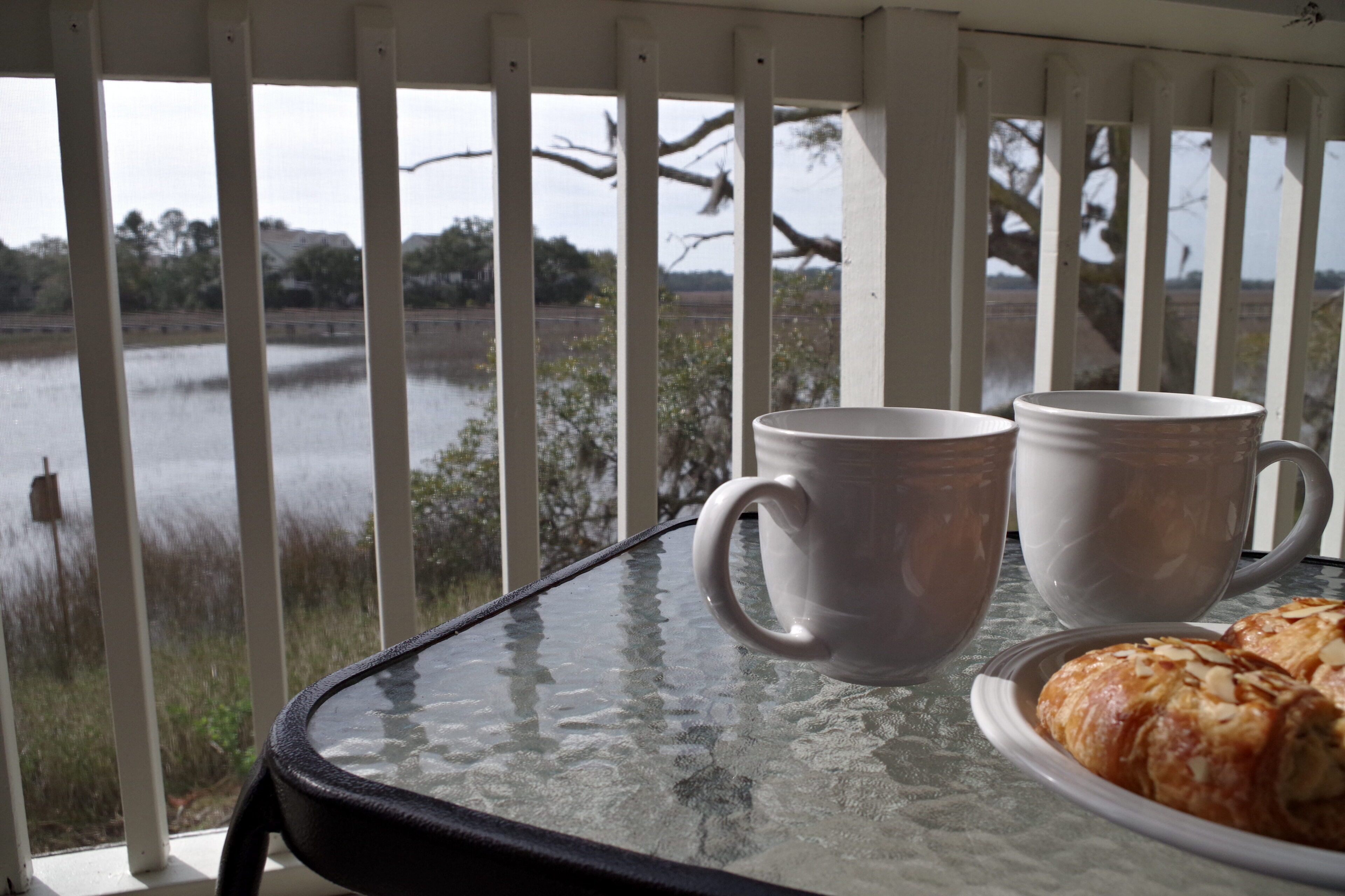 A glass table with two white mugs and a plate of pastries, set against a view of a lake and trees.