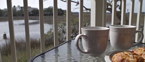 A glass table with two white mugs and a plate of pastries, set against a view of a lake and trees.
