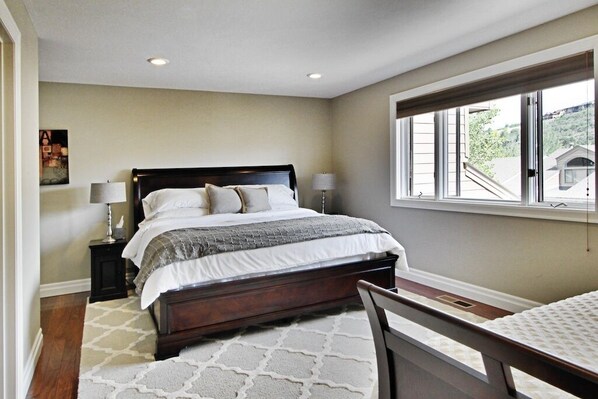 A bedroom with a large bed, two bedside tables with lamps, a window with a view of a house, and a patterned rug.